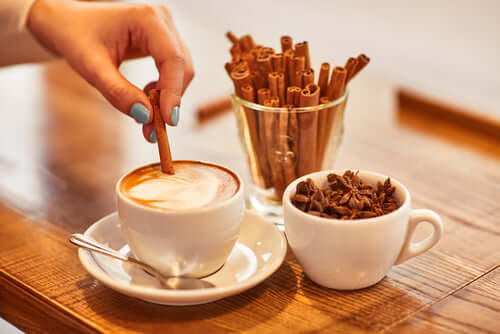 A hand stirring a cinnamon stick in a cup of coffee beside a bowl of coffee beans and cinnamon sticks.